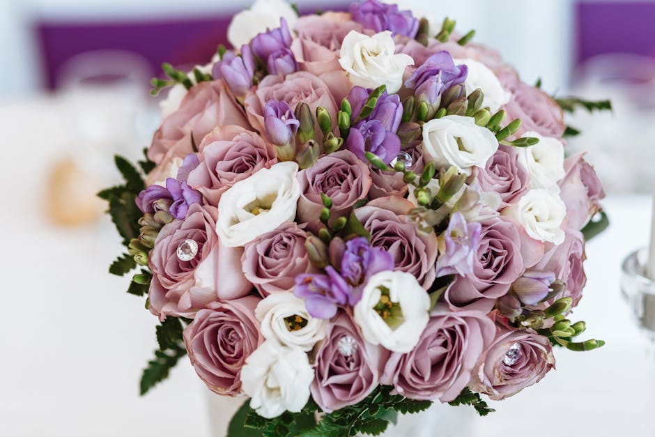 A close-up of a floral bouquet arranged with pink roses, white eustoma, and purple freesia, accented with small green leaves and decorative clear rhinestones. The flowers are fresh, with softly textur