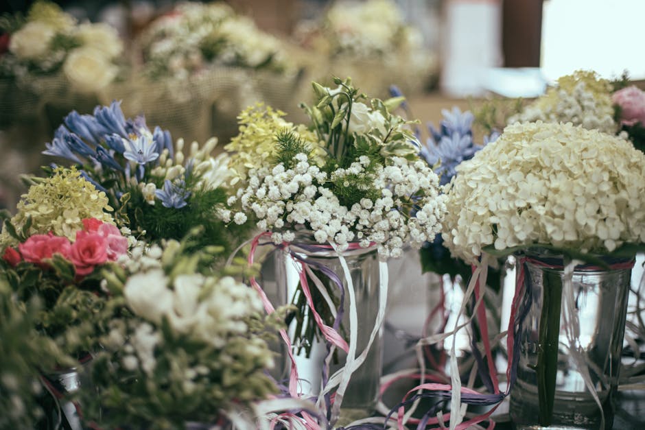 A collection of floral arrangements featuring white hydrangeas, pink roses, blue irises, and delicate baby's breath, all displayed in clear glass vases decorated with pink and white ribbons. The flowe