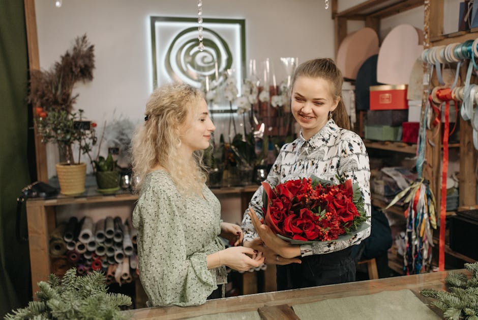 A young woman with light brown hair tied back, smiling as she receives a vibrant floral bouquet from a florist in a shop. The bouquet features a lush arrangement of red roses, red anthuriums, and smal