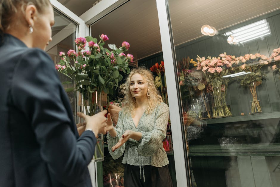 A young woman with curly blonde hair and light skin stands inside a flower shop, smiling as she receives a tall bouquet of pink roses wrapped in brown craft paper from a florist. The bouquet features 