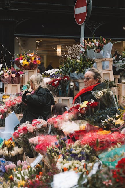 A vibrant outdoor flower market under a dark canopy, featuring an array of fresh, colorful floral arrangements and bouquets arranged on tables and displayed in wooden crates. The flowers include red a