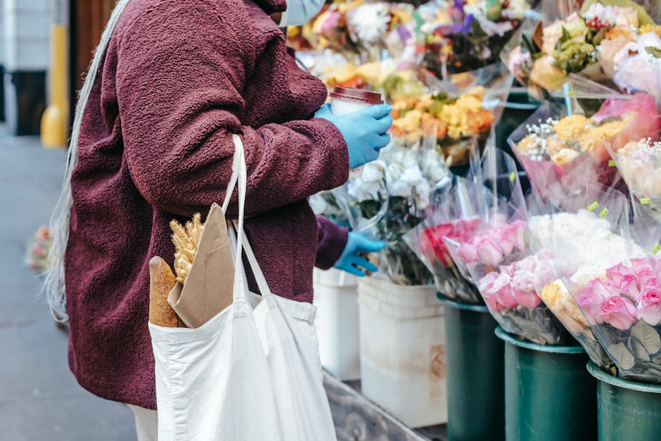 A person wearing a burgundy fleece jacket and light blue gloves is selecting a bouquet of fresh flowers at an outdoor flower stall. They are holding a takeaway coffee cup in one hand and carrying a ca