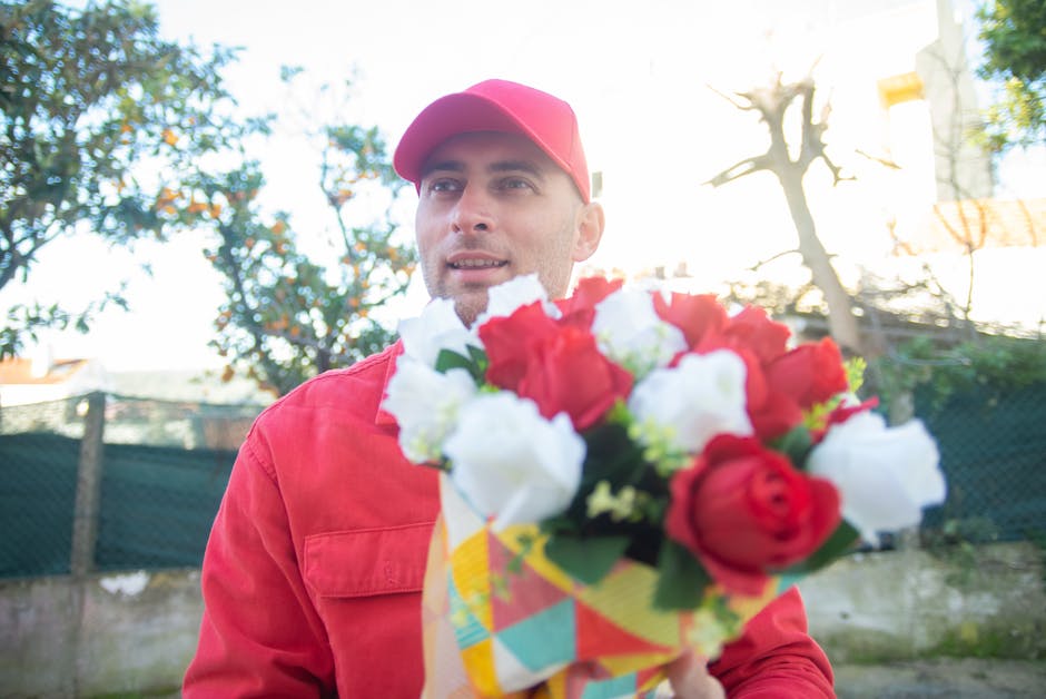 A man dressed in a red jacket and matching red cap is holding a colorful bouquet of fresh flowers, including red and white roses, wrapped in patterned paper. He is outdoors in a sunlit garden with tre