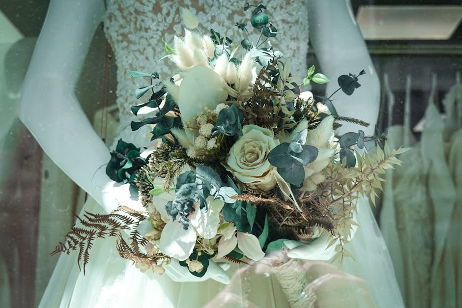 A bouquet arrangement held by a person in wedding attire, featuring a mix of white and cream roses, eucalyptus leaves, fern fronds, and dried brown foliage. The flowers are fresh with soft, velvety pe