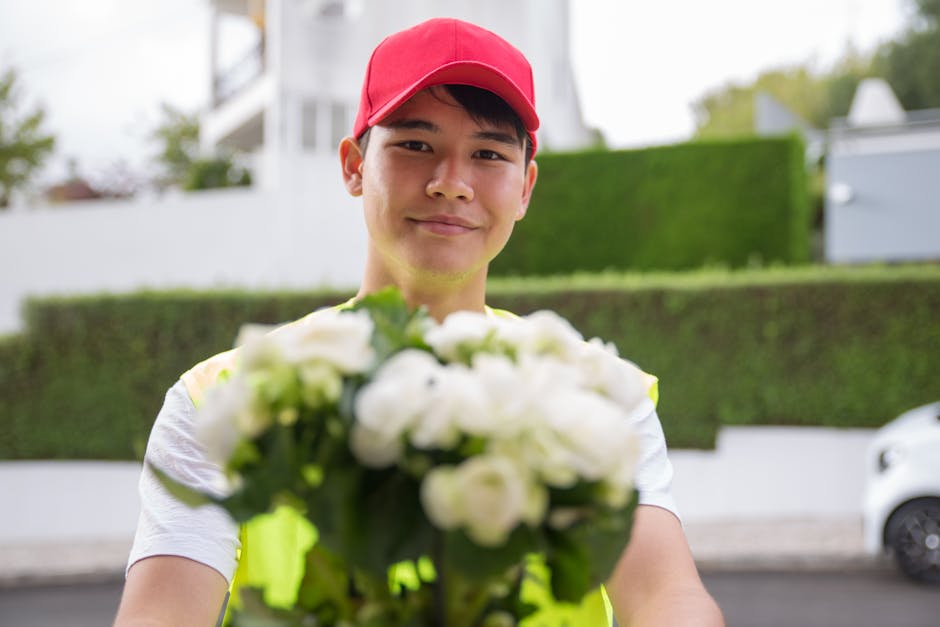 A delivery person wearing a red cap and a yellow reflective vest is holding a bouquet of fresh white roses and green foliage, extending it towards the camera. The flowers appear vibrant and well-hydra