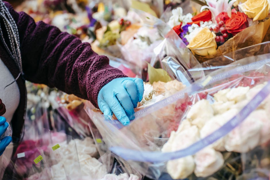 A florist wearing a dark purple velvet jacket and a gray top is seen selecting a floral arrangement wrapped in clear plastic from a display of various freshly cut flowers. Their hand, covered with a b
